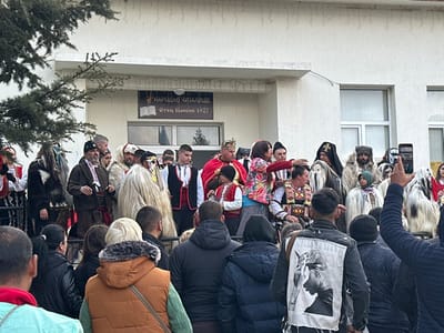 The image captures an outdoor cultural event in Novo Panicharevo, Bulgaria, specifically in front of the "НАРОДНО ЧИТАЛИЩЕ Отец Паисий 1927" (National Cultural Center Father Paisiy 1927) building. In the midground, approximately twenty individuals are gathered on a low platform or stage. This group consists of performers and participants dressed in various traditional Bulgarian costumes, including several individuals in shaggy white and dark fur Kukeri masks and full-body outfits. A man wearing a red cloak and a golden crown stands centrally, alongside a woman in a colorful floral jacket who is actively gesturing with her right arm. Nearby are a child in a traditional white shirt and red vest, and a man in a dark robe with a cross on his headwear, resembling an Orthodox priest. The foreground is occupied by a crowd of spectators, largely viewed from behind, observing the event. One person on the far right holds a smartphone vertically, recording the scene. The background features the light-colored, two-story community building with two large windows, partially obscured by a tree branch on the left. The sky is overcast.