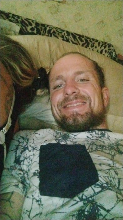 The specific location within the city of Mariposa, United States, is not discernible from the image; it appears to be an indoor residential setting.
The scene features two individuals positioned closely, likely lying down. The primary subject is a male adult with light skin, a full beard, and a ring piercing in his left nostril. He is smiling broadly, revealing teeth, and looking directly towards the camera. He wears a light-colored t-shirt with a dark, abstract, or camouflage-like pattern and a prominent solid black rectangular patch on the chest. To the left of the male subject, a second individual's long, light-colored hair (blonde or light brown) is visible, along with a portion of their left shoulder and a thin white strap, possibly from a bra or tank top. The second individual's face is not visible.
The environment appears to be a bedroom or a comfortable lounge area. Visible objects include a light-colored pillow behind the male subject's head and a distinct black and tan leopard-print fabric, likely a blanket or bedding, in the upper portion of the frame. The lighting is soft and even, consistent with indoor ambient light. No specific actions or interactions beyond their shared proximity and the male's direct gaze at the camera are evident.