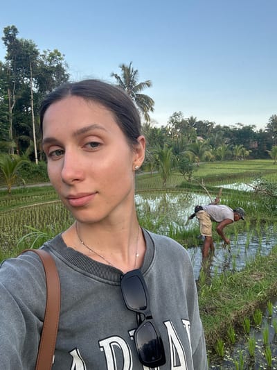 A woman takes a selfie in the foreground, with a rice paddy and a farmer in the background. The woman has dark brown hair and is wearing a grey sweatshirt and a brown bag. She has a necklace with small charms. Sunglasses are hanging from her sweatshirt. The rice paddy is lush and green, with water reflecting the sky. A man, presumably a farmer, is bent over, tending to the rice plants in the shallow water. The scene appears to be in a rural setting, likely Tetebatu, Indonesia, with palm trees and other foliage visible in the distance. The sky is a clear, pale blue.