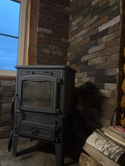 This image captures the interior of a room, likely a home in Shebarshino, Russia, featuring a dark grey wood-burning stove positioned in the corner. The stove has a glass door to view the fire and intricate decorative details. The walls are constructed of dark, textured brickwork, varying in shades of brown and grey. To the left of the stove, a large window with a white frame is visible, letting in a blueish light. Adjacent to the stove, a stack of firewood, including birch logs with distinctive white bark, rests on the floor. A metal grate or rack is partially visible to the right of the firewood, and a red box with Russian text, possibly containing matches, sits atop the logs. The floor appears to be a dark, tiled surface. The overall ambiance is rustic and cozy, suggesting a functional and decorative element for warmth within the dwelling.