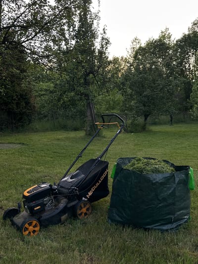 This image depicts a residential outdoor scene in Staro Selo, Bulgaria, likely during late afternoon or early evening under a slightly overcast sky. The foreground is dominated by a black and orange McCulloch brand gasoline-powered lawnmower, positioned on a grassy lawn. The lawnmower features orange wheels and visible engine components. Adjacent to the lawnmower, a large, dark green woven polypropylene bag, filled to capacity with freshly cut green grass, rests on the ground. The bag has bright green handles on its sides. The immediate grassy area appears recently mowed, indicating recent lawn maintenance activity. The background features a dense collection of various trees with rich green foliage, suggesting a rural or suburban garden environment. Some trees in the upper left section show small, rounded shapes among their leaves, potentially indicating fruit trees. Further back, denser vegetation and taller grasses are visible, bordering the maintained lawn area. No individuals are present in the scene.
