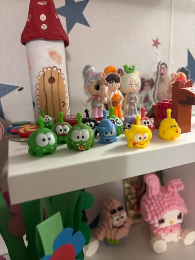 The image depicts a collection of toys and decorative items arranged on two white shelves within an indoor setting, consistent with a child's room in Krasny Klyuch, Russia.
On the upper shelf, a primary grouping of small plastic figurines, predominantly green, blue, yellow, black, and purple "Om Nom" characters from the "Cut the Rope" game, are displayed. A red and white circular lollipop-shaped object is to the far left of these figures. To the right, a structure composed of brown and red interlocking plastic bricks, resembling LEGO, is partially visible. Standing prominently on the far left of this shelf is a handcrafted cylindrical decoration with a red pointed roof, resembling a mushroom or fairy house, featuring a drawn brown door. In the background, behind the Om Nom figures, a white wall is decorated with multiple blue star decals and one small red star decal. Several additional figurines and cardboard cutouts are visible on a surface against this wall, including a "LOL Surprise!" doll with glittery silver hair, a male character in orange overalls holding blueprints, another light-colored character with a green alien on its head, and two human-like cardboard cutouts, one with a beard.
The lower shelf, partially visible beneath the upper one, features green paper or felt cutouts resembling foliage and colorful paper flower cutouts in red and blue. Displayed on this lower shelf are two block-style figurines: a pink and white "My Melody" character on the right, and a peach-colored "Patrick Star" character on the left. All subjects are static, arranged for display.