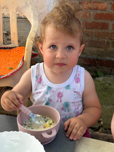 A young child with blond curly hair and blue eyes is sitting at a table outdoors and eating. The child is wearing a white sleeveless shirt with pink and blue bunny patterns. They are holding a pink spoon that is partially in a pink bowl of food, which appears to be a soup or cold dish with visible ingredients like potatoes and herbs. To the left of the bowl, there is a white bowl with a scalloped edge. The background includes a brick wall and a portion of a yellow plastic chair with an orange and green woven mat. The overall scene suggests a casual meal, possibly in a backyard or garden setting in Komsomolskiy, Russia.