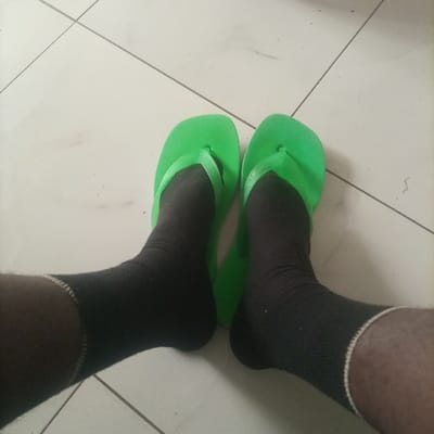 The image depicts a close-up, downward view of two lower legs and feet, located in Kimorori/Wempa ward, Kenya. The individual's feet are clad in black ankle-height socks, each featuring a white band at the top cuff. Over these socks, a pair of bright green flip-flops are worn, positioned on a light-colored tiled floor. The floor tiles are square or rectangular with visible dark grey grout lines, indicating an indoor setting. Minor surface debris or scuff marks are present on the tile surface. No other individuals or distinguishing features of the wider environment are visible.