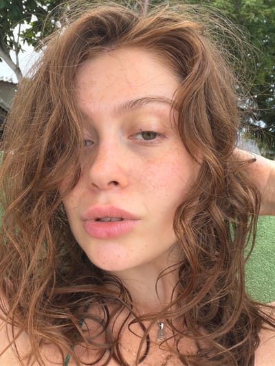 This is a close-up, rotated, outdoor photograph of a young person's face. The subject has light skin with freckles, light brown wavy hair, and green eyes. Their lips are slightly parted, revealing their teeth. The person is lying down, with their head tilted and their hair spread out. A green textured surface, likely a towel or cushion, is visible behind them. Trees and foliage are also visible in the background, suggesting an outdoor setting during the day. The overall mood is relaxed and natural. There is no visible text in the image.