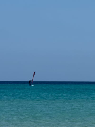 The image displays a lone windsurfer navigating a vast expanse of water under a clear sky. The composition is vertically oriented, with a smooth, light blue sky occupying approximately two-thirds of the frame on the left, and a body of water taking up the remaining right third. A sharp, vertical line acts as the horizon, cleanly separating the sky from the water.
The water, on the right side, is a vibrant teal and turquoise, showing subtle textures of ripples or small waves across its surface, suggesting a gentle breeze. The sky is a uniform, cloudless light blue, indicating bright, sunny weather.
Centrally located along the vertical horizon line is the windsurfer. The individual is largely silhouetted in dark tones, actively engaged with a windsurfing board and sail. The sail is a prominent feature, colored a distinctive red or maroon, and appears to be catching the wind. The person is leaning into the sail, demonstrating movement and control, though specific facial features or expressions are not discernible. The overall impression is one of serene, solitary recreation.
The scene suggests a warm, bright day, ideal for watersports. There are no other objects, boats, or land visible, emphasizing the open nature of the sea and sky. No visible text is present in the image. The location context of Pájara, Spain, aligns with this coastal activity.