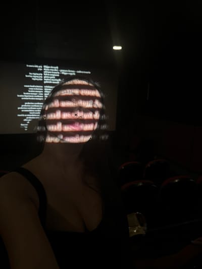 The image depicts a person indoors in a dimly lit environment. The person's face is illuminated by a projection consisting of vertical bars of light and dark stripes, creating a grid-like pattern. Adjacent to the face is a display of text, also illuminated by the projection. The background is largely dark, with some indistinct objects and surfaces barely visible. The location, Lesnoi Gorodok, Russia, is not visually evident within the image.