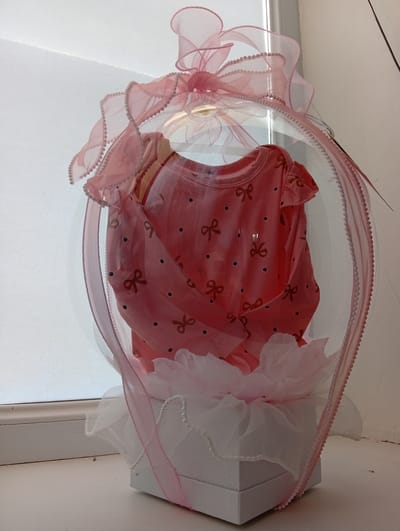 A pink baby onesie with a bow pattern and black polka dots is displayed inside a clear plastic sphere. The sphere is adorned with pink ribbon and pearls. The sphere sits atop a white box, which is decorated with white and pink tulle and more pearls. The entire arrangement is placed on a white surface, possibly a windowsill, against a background of a lightly textured wall and a window with a translucent blind. The image is taken from a slightly low angle, looking up at the display. There are no people or animals present in the image. The location is specified as Tomai, Moldova.