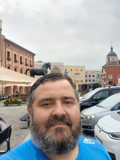The media file is a vertical, low-angle selfie of a man with a beard, wearing a blue t-shirt with the "adidas" logo visible on the sleeve. The man is in the foreground, with his face filling a significant portion of the frame. Behind him, the scene depicts an outdoor setting with several cars parked, including a white electric car. In the background, there are buildings with colorful facades, suggesting an urban or town environment. One building has a prominent clock tower. A statue of a black animal, possibly a dog or a panther, is visible on a pedestal in front of one of the buildings. The sky is overcast, indicating a cloudy day. The overall impression is a casual snapshot taken during the daytime, likely in a European town square or street given the architecture and parked vehicles. The man's expression is neutral. Visible text includes the "adidas" logo on his shirt and a license plate on one of the cars. The presence of the clock tower and the style of the buildings suggest the location could be in Germany.