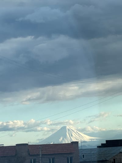 An eye-level, vertical shot, taken in Verin Ptghni, Armenia, shows a cloudy sky to the left with power lines running across it, while a mountain can be seen on the right. Building structures are visible in the lower right corner of the frame. The mountain's peak is snow-capped and contrasts against a sky with scattered clouds.