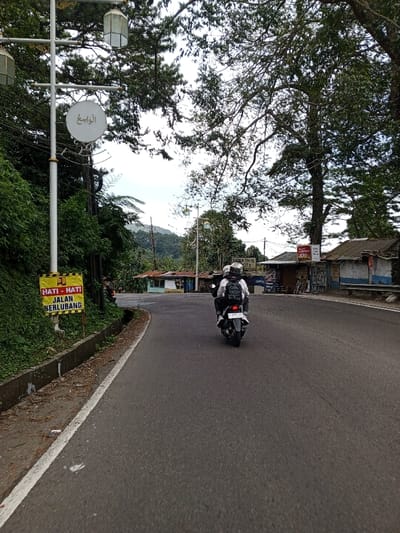 The image depicts a two-lane asphalt road in Ciloto, Indonesia, curving to the right under an overcast sky. A white scooter-type motorcycle, bearing license plate B 5372 JY, travels away from the viewer in the center of the right lane. It carries two individuals wearing helmets; the person in the rear wears a white helmet and a light-colored top, and the driver wears a darker top with a backpack and a white helmet.
To the left of the road, a concrete drainage ditch runs alongside a steep, heavily vegetated embankment. A yellow warning sign with red text, partially obscured, reads "HATI - HATI JALAN BERLUBANG" (Caution - Potholed Road). A tall, light-colored streetlamp post stands near the sign, featuring three lantern-style light fixtures and a circular white sign bearing Arabic script ("الواسع" - Al-Wasi').
To the right of the road, several small, basic structures with corrugated metal roofs line the roadside, appearing to be shops or stalls. Signs are visible on these structures, including one red sign with white text and another, partially visible, that reads "WISMA TITAN JAYA FOTO". A wooden bench is positioned in front of one structure. Large, mature trees with dense green foliage canopy over parts of the road and structures. In the background, green hills and mountains are visible under a hazy sky.