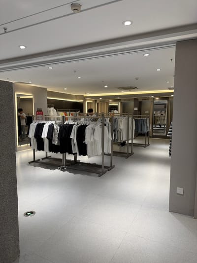 A retail store located in Qinhuai District, China, displays men's clothing on racks. The store features a bright, modern interior with white tiled floors and a suspended ceiling with recessed lighting and ventilation. Clothing racks are arranged in the center of the store, showcasing a variety of t-shirts and other garments in white, black, and blue denim. Mirrors are strategically placed throughout the store, including a large mirror on the left wall reflecting a man in the process of selecting clothing. In the background, several fitting rooms with beige curtains are visible, along with additional clothing racks. A green directional arrow illuminated on the floor indicates an exit or pathway. Electrical outlets are visible on a wall to the right of the frame.