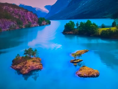 The image depicts a serene landscape of a large body of water surrounded by mountains and islands. The water is a vivid blue. On the left side of the frame, a mountain with exposed rock and green vegetation rises, with a road visible along its base. In the background, snow-capped mountains can be seen. The right side of the frame shows darker mountains descending to the water's edge. Several small, tree-covered islands dot the water. The islands vary in size and shape, some with a single tree or a small cluster of trees.
This image is most likely from somewhere in Norway, as this is a very typical landscape from the Nordic country.
There is no evidence that this image is related to Basingstoke and Deane, United Kingdom.