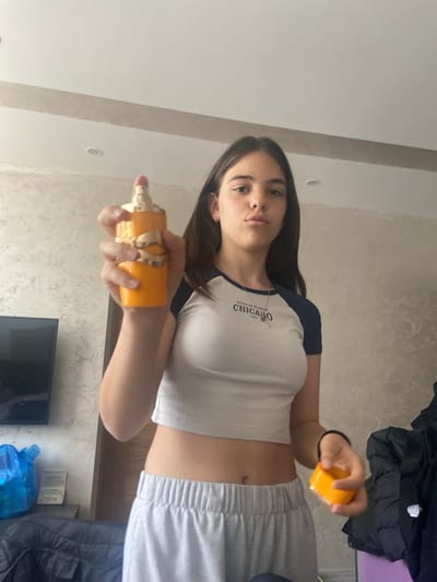 A teenage girl is indoors, holding a yellow perfume bottle in her left hand and a yellow object in her right. She is wearing a white and navy blue top with the word "CHICAGO" on it and grey sweatpants. The background includes a patterned wall, a TV, and possibly clothing. There is no confirmed evidence to associate this picture with the city of Dragovishtitsa, Bulgaria, and therefore I can not provide location details.