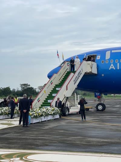 An airport tarmac scene captures a formal arrival or departure ceremony under an overcast sky. A large blue passenger aircraft, with "ITALIA" partially visible on its fuselage, is parked with its door open. A set of mobile stairs, marked "FLOTA AÉREA PRESIDENCIAL" in Spanish, connects the aircraft to the ground.

At the top of the stairs stands a prominent figure dressed in white robes, consistent with the Pope or a high-ranking religious leader, accompanied by individuals in dark suits and one in a naval uniform. On the side of the stairs, flags of Vatican City and Equatorial Guinea are displayed, along with small signs partially visible that read "REPUBLICA DE GUINEA ECUATORIAL."

Extensive arrangements of white and yellow flowers line the base of the stairs and extend along the tarmac. Several people in formal attire, including media personnel with cameras, are gathered on the ground, observing the activity. Aircraft landing gear and ground support equipment, including one labeled "AIR BUS 340," are visible. The tarmac appears damp, suggesting recent rain or high humidity. In the background, lush green trees rise under the cloudy sky, characteristic of a tropical location like Malabo, Equatorial Guinea, where the event is taking place. The overall impression is one of a dignified and ceremonial occasion.