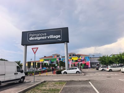A wide shot shows the Palmanova Designer Village with a bright, cloudy sky overhead. The architecture of the village is modern and features vibrant, geometric murals in bold colors like pink, yellow, blue, and green. Several trees line the area in front of the buildings. In the foreground, a white delivery van is parked on the left, and a white hatchback car is parked on the right. Parking lines are clearly marked on the asphalt. A yield sign is prominently displayed on a pole near the center of the frame. A person in a blue shirt walks away from the camera in the middle distance, towards the village buildings. The scene appears to be during daylight hours. The location is identified as Palmanova Designer Village, and the text "FREY" is visible on the large signage.