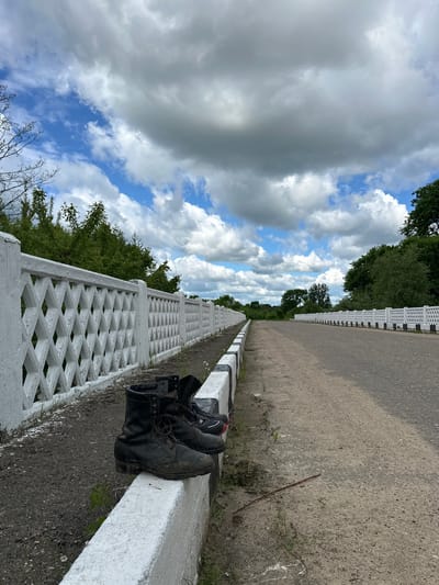The image shows a pair of black boots placed on a white and black patterned concrete fence. The fence runs alongside a grey asphalt road that stretches into the distance. Trees and foliage line the other side of the fence. Above, the sky is filled with clouds, some white and fluffy, others darker and grey, with patches of blue sky visible. The location is in Валожынскі сельскі Савет, Belarus.
