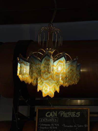 An ornate, illuminated chandelier with a gold-colored metal frame and multiple tiers of beaded fringes dominates the center of a dimly lit interior in Montuïri, Spain. The chandelier's fringes are predominantly light blue/teal and yellow, forming intricate patterns, and are composed of numerous small beads. Several internal light sources within the chandelier are active, casting a warm glow. In the upper background to the left, large, dark brown, cylindrical objects, resembling wine or oil barrels, are partially visible, one featuring a dark strap. In the lower right foreground, a partially visible chalkboard menu displays "CAN PIERES" at the top, followed by "ENTRANTES:" (Appetizers) and handwritten items in Spanish, including "Calamarés 16'90€", "Croquetas 12'50€", and "Pimientos de Padrón 10€". No individuals or discernible actions are present within the frame.
