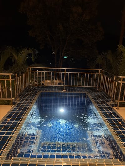 This image depicts a small, tiled swimming pool at night. The pool is illuminated by a single underwater light, casting a bright glow on the blue water and creating ripples. The pool is surrounded by a stainless steel railing. In the background, palm trees and a large tree are visible against a dark sky, with some distant lights suggesting an urban or suburban setting in Ban Kao Dua, Thailand. A white car is partially visible behind the railing, parked in an unseen area. The overall scene is tranquil and suggestive of a private or resort setting.