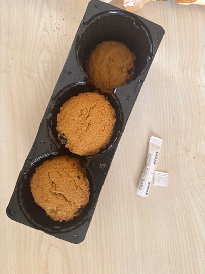 In Aragats, Armenia, a forensic examination of the scene reveals a top-down view of three round, light brown cookies, each containing dark inclusions, resting in separate compartments within a black plastic tray. The tray is positioned diagonally from the lower left to the upper right on a light-colored wooden surface. Adjacent to the right side of the tray, a white paper sugar packet, marked with "SUGAR" and "CAXAP" (Cyrillic for sugar), lies partially torn open. Fine crumbs are scattered across the wooden surface, particularly around the upper portion and near the tray. A crumpled, out-of-focus piece of light-colored packaging is partially visible in the top right corner of the frame.