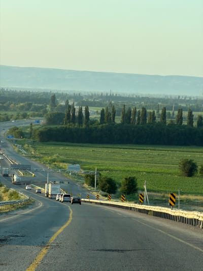 The image depicts a wide, paved highway curving through a rural landscape with agricultural fields and scattered trees under a pale, hazy sky. The road, marked with a yellow center line, has multiple lanes and is bordered by a metal guardrail and yellow and black hazard signs. Several vehicles, including trucks and cars, are visible on the highway, traveling in both directions. The surrounding terrain is characterized by flat, green fields, with rows of tall, slender trees forming a line in the mid-ground. In the distance, rolling hills are faintly visible through the haze. The overall impression is one of a highway traversing a vast agricultural region during daylight, possibly in the early morning or late afternoon given the soft light. There is no indication of the city of Mukry, Kazakhstan, in this image.