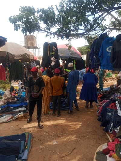 An outdoor market scene in Zangon Aya, Nigeria, displays multiple individuals amidst clothing stalls under natural daylight. The ground is dirt. In the foreground, folded clothing is piled on tarps on either side of a central dirt path. A male figure wearing a black patterned shirt, black pants, red baseball cap, and dark slides stands centrally, holding a small burgundy bag. To his left, a market stall features a beige umbrella canopy with clothes hanging from it, and a person is crouched near piles of garments on a blue tarp. To his right, a cluster of male figures, mostly with their backs to the camera, are observed: one in a yellow striped garment, one in an orange shirt and red cap, and another in a light blue traditional robe and white cap, all appearing to browse or interact with goods. Further right, a figure in a dark blue robe stands facing away. Overhead, various articles of clothing, including a black hoodie with "KEN" in yellow text and a blue hoodie, are suspended from lines or tree branches. In the background, a large, rusty metal water tank on a tall stand is visible beneath a clear sky and scattered green foliage.