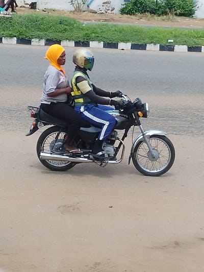 In Oja Oba, Oko Erin, Nigeria, two individuals are riding on a motorcycle. The person in the front is wearing a yellow headscarf and a grey t-shirt with the number "20" visible on it. The person in the back is wearing a helmet with a reflective visor and a yellow and green vest over a black garment. They are both wearing dark pants. The motorcycle is black with chrome accents, including the exhaust pipe and handlebars. The scene takes place on a paved road with a grass embankment and black and white striped border in the background.