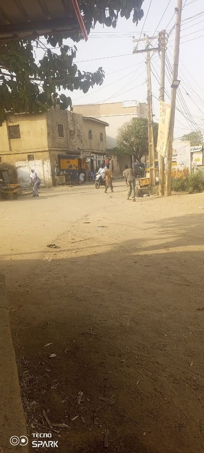 The image depicts an unpaved street scene in Tudun Wazurchi, Nigeria, under a hazy daylight sky. The foreground and mid-ground consist of dry, reddish-brown earth with scattered debris including small pieces of wood.

On the left side of the frame, a two-story building with a light beige facade and multiple windows stands at a corner. Adjacent to it, a single-story structure features a commercial storefront with a closed yellow or orange roller shutter, flanked by dark blue panels. Several individuals are visible near this storefront; at least two men are seated, and one man stands facing the storefront. To the far left, a man in a white shirt and grey trousers stands next to a two-wheeled wooden cart laden with produce.

In the mid-ground, approximately eight to ten individuals are present. A woman wearing a light grey hijab and dress walks from right to left. A man in a light brown tunic and dark trousers walks from left to right, positioned slightly behind a motorcycle with a rider. Another man, wearing a light-colored top and jeans, walks further right, appearing to adjust something or having an uneven stride.

On the right side of the image, multiple wooden utility poles rise, supporting a dense network of overhead electrical wires and a single street light fixture. A vertical banner, featuring an illustrated man in a red cap and text, is attached to one of these poles. Beyond the poles, another vehicle, possibly a tricycle, is partially visible. Further back, another multi-story building with a lighter facade and a flat roof is discernible.

Tree branches with green foliage extend into the upper left quadrant, providing partial shade. A smaller, less leafy tree is visible in the middle ground, near the center-right. Shadows are cast on the ground from the left side of the frame, indicating the position of the sun.