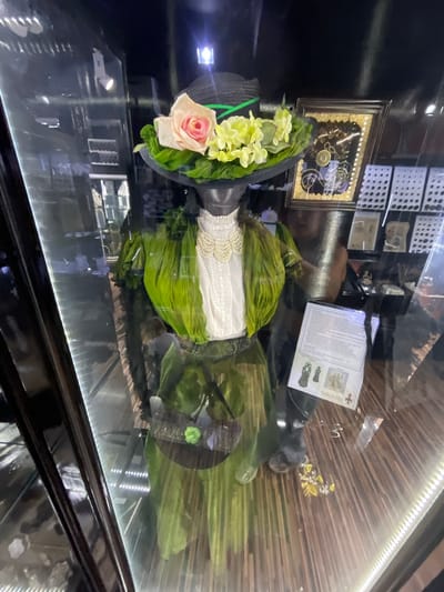 An interior shot shows a mannequin dressed in an antique green dress with layered fabric and a white lace collar. The mannequin wears a black hat adorned with a pink rose, yellow flowers, and green leaves. A document is displayed in front of the mannequin on a stand. The background reveals a museum display with framed items and shelves. The scene is lit with artificial lights, reflecting on the glass case.