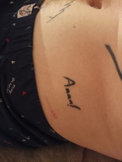 The image shows a close-up view of a person's arm and leg, with a dark blue fabric with a pattern of small sailboats and red and white triangles. There is a visible tattoo on the arm that appears to read "Aaaaj!". Another tattoo, a thin black line, is partially visible on the upper arm. The skin on the arm has some minor redness, suggesting recent irritation or a fresh tattoo. The lighting suggests an indoor setting or possibly overcast weather. The visible text on the fabric is difficult to decipher completely, but "black sea" is partially legible. The context indicates the location is Zapadny, Russia.