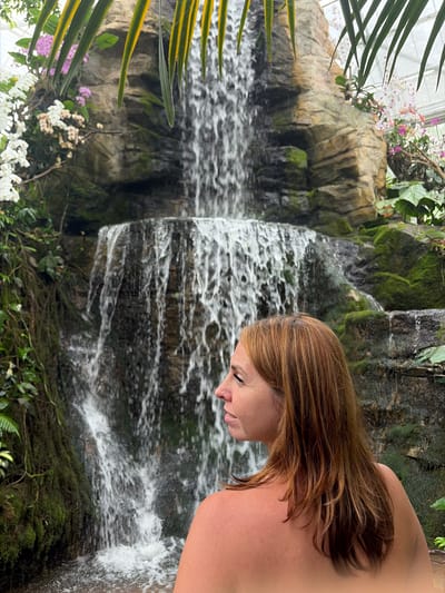 The image depicts a woman with shoulder-length, auburn hair, viewed from the back and right side of her face, standing in front of a cascading waterfall. She is shirtless, with her skin appearing tanned. The waterfall is set within a lush, green environment, featuring moss-covered rocks and various plants, including orchids with white, pink, and purple blooms. Large, elongated green leaves with yellow stripes overhang the top of the frame, partially obscuring the waterfall and rock formation. The background suggests an indoor setting, possibly a botanical garden or greenhouse, indicated by the light filtering from above. The location is identified as Pong Yaeng, Thailand.