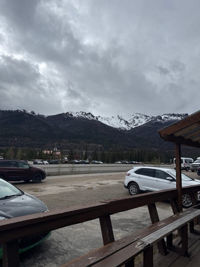 The image captures a parking lot scene in Healy, United States, with a mountain in the background, partially covered in snow. Overcast skies fill the upper left portion of the frame. A line of parked cars is visible along the right side of the lot, with various car models. The scene is viewed through the structure of a wooden railing, which frames the parking lot.