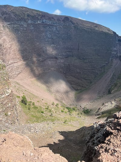 The image shows a view into the crater of Mount Vesuvius, located near Ercolano, Italy. The crater is a large, bowl-shaped depression with steep, rocky sides. The interior of the crater is covered in scree and sparse vegetation, with a visible path winding down into the depths. The sky above is a clear blue with scattered white clouds. Shadows from the crater rim and a cloud are cast across the volcanic landscape, highlighting the varied textures of the rock and soil. The foreground consists of rough, reddish-brown volcanic rock, suggesting the viewpoint is from the edge of the crater rim.