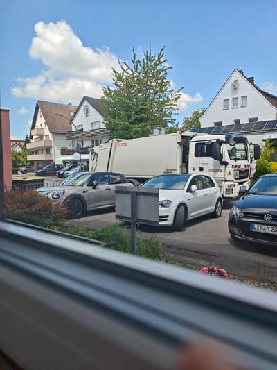 In Bad Meinberg, Germany, a refuse collection vehicle is parked on a street adjacent to residential buildings. The truck is white with a large, beige refuse body. A white passenger car and a silver hatchback are parked in front of the refuse truck. The buildings in the background feature red-tiled roofs and balconies. The sky is blue with scattered white clouds. A tree with green foliage is visible between the buildings.
