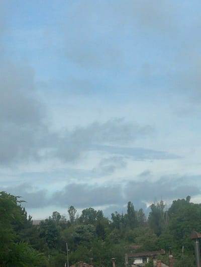 The image displays a landscape scene, primarily featuring an overcast sky above a dense line of trees and foliage. The image is oriented vertically, showing the sky on the left and the treeline on the right when viewed as rotated 90 degrees counter-clockwise.

The left two-thirds of the image (when correctly oriented) are dominated by a pale blue-grey sky, filled with soft, diffused clouds. The clouds appear as a continuous, somewhat textured blanket, indicating an overcast or heavily cloudy day. There are no clear breaks in the cloud cover or direct sunlight visible, suggesting soft, ambient light.

The right third of the image (when correctly oriented) is occupied by a lush, green treeline and dense foliage, stretching across the entire vertical length of the frame. The trees are a mixture of various types, appearing full and healthy. Within and behind the trees, several indistinct structures or elements are visible. Towards the top right, what appear to be parts of buildings or structures, possibly with reddish-brown rooftops or weathered surfaces, peek through the canopy. Further down, thinner, possibly deciduous trees with fewer leaves or distant structures are faintly silhouetted against the sky. Vertical poles, possibly utility poles, are also discernible within the dense vegetation.

No people or discernible activities are taking place. The scene captures a static natural landscape with hints of human presence in the background. The time of day appears to be daytime, likely morning or afternoon, given the ambient light, but definitely not bright midday due to the heavy cloud cover. The weather is cloudy and overcast. Given the provided context, this scene depicts an area in Yana, Bulgaria. There is no visible text in the image.