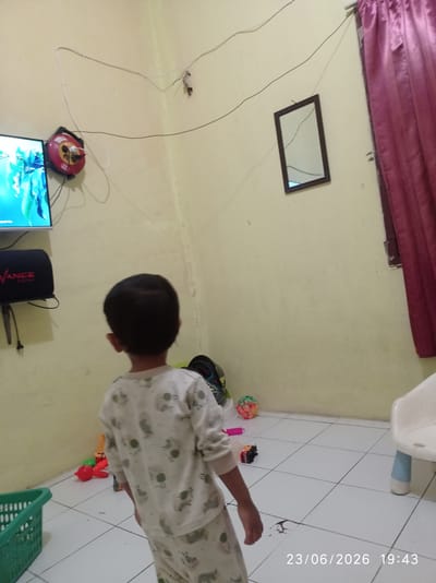 In a dimly lit room, a young child wearing pajamas is standing facing away from the camera, looking towards a television screen. The room has light yellow walls and a tiled floor. To the left of the child, a television is mounted on the wall, displaying a blue and green image. Below the television is a black speaker with red lettering that says "VANCE".

On the wall above the television, there is a red circular device with a white cord plugged into it, attached to a white wire that runs across the wall. A small, dark object is also attached to the wire near the top right of the frame.

To the right of the child, there is a small mirror with a dark frame hanging on the wall, and a dark red curtain partially visible on the far right. The floor is scattered with various children's toys, including a green basket in the bottom left corner, a red ball, and toy cars. A white child's chair with blue legs is also visible on the right side of the frame. The timestamp "23/06/2026 19:43" is overlaid on the bottom right of the image. This scene is likely from a residential dwelling in Sukaraja, Indonesia, given the context.