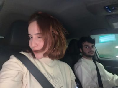 Two people are in a car. The person on the left has dark hair and a beard and is wearing a white shirt with a black strap. The person on the right has reddish-brown hair and is wearing a tan sweatshirt with a black strap. The location appears to be inside the car. There is no indication of Grigalati, Georgia.