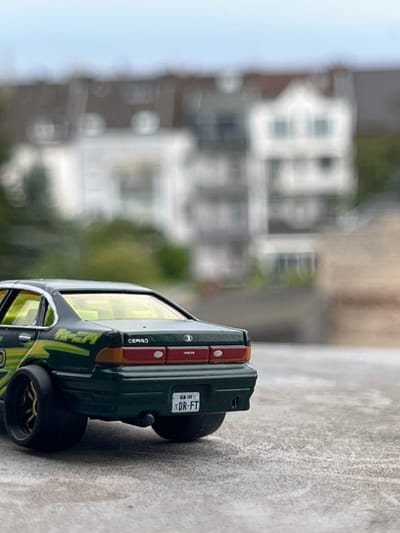 The media file shows a close-up, rotated view of a green toy car with yellow and white racing stripes. The car has detailed rear lights, a license plate that reads "DR-FT," and a "CEPTO" label on the side. The background is blurred, showing a street scene with buildings and greenery, suggesting an outdoor setting. The overall impression is that of a hobbyist displaying their toy car collection. The image's orientation and the blurred background suggest it was taken outdoors, possibly in a residential area or a park. No specific time of day or weather conditions can be determined from the image. The location context provided is Neuss, Germany, but there are no visible cues within the image itself to confirm this.