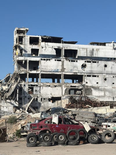 In Jabalia, Palestinian Territories, a multi-story concrete building stands severely damaged, with large sections of its facade missing and rubble accumulated at its base. The building's interior floors are exposed, and some sections show signs of fire damage. In the foreground, several destroyed or dismantled vehicles are piled up, including a red car with its doors open and multiple tires scattered around it. The environment appears to be an urban area that has experienced significant destruction.