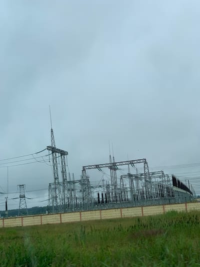 The image shows an outdoor electrical substation with numerous metal towers, complex electrical equipment, and high-voltage power lines under a cloudy sky. In the foreground, there is a low concrete wall with a textured pattern, and beyond that, green grass and vegetation. The substation is situated in an open area, with a hill and some trees visible in the background. The overall scene suggests an industrial landscape in the Курапольскі сельскі Савет region of Belarus. There are no people or animals visible in the image.