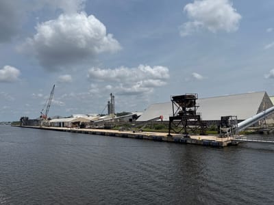 This is a medium-shot, eye-level, full exterior, daytime, horizontal, color photograph of the Port of Brunswick, Georgia, USA. The image captures a long, industrial waterfront scene. In the foreground, a dark, rippling body of water occupies the lower half of the frame. Along the shore, a concrete pier runs horizontally across the image.
The pier is lined with various industrial structures. On the far left, a large construction crane with a dark boom stands tall. Adjacent to it are several large, dark, rectangular buildings. Further to the right, a series of large, white, A-frame industrial buildings with metal roofs dominate the middle ground. Conveyor belts, some elevated on metal structures, are visible extending from these buildings towards the water. A prominent, dark metal industrial gantry with complex machinery is situated in the right-center of the frame, overlooking a wide conveyor belt that leads to a large, white structure. Yellow bollards are intermittently placed along the edge of the pier. The sky above is partly cloudy with large, puffy white and grey clouds scattered against a pale blue background. There are no people visible in the image. The scene depicts a working port with various material handling and storage facilities.
