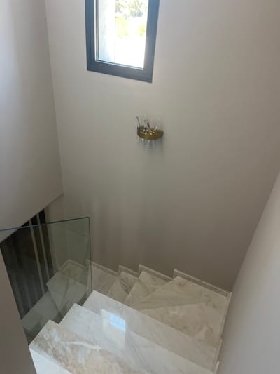 A modern interior staircase with marble steps and a glass railing is captured from a high angle in the Moni Community, Cyprus. The walls are painted a light, neutral color. A small, decorative wall sconce with crystal elements is mounted on the right wall. A window with a dark frame is positioned high on the left wall, allowing natural light to illuminate the scene. The overall impression is one of clean lines and sophisticated design.