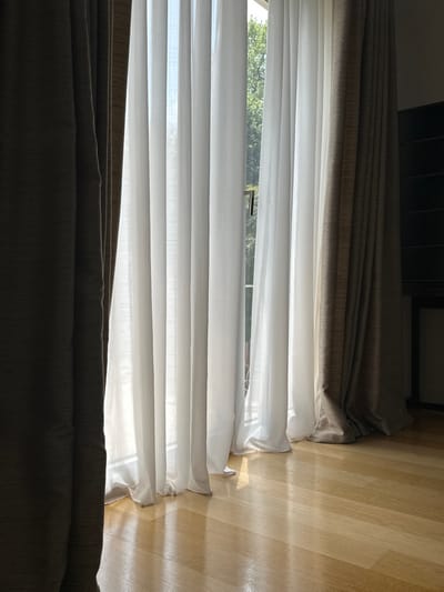The image displays an interior room located in Vandœuvres, Switzerland, focused on a large window or glass door. The floor consists of light-toned, polished hardwood, reflecting incoming natural light. Two layers of curtains are present: an inner layer of multiple vertical white, translucent sheer panels fully drawn across the window, and an outer layer of heavier, opaque drapes, dark brown or greyish-brown, partially drawn open with one panel visible on the far left and another on the far right. Bright sunlight filters through the sheer curtains, illuminating the room. Outside the window, obscured by the sheer fabric, green foliage from trees or bushes and a bright sky are discernible. In the background on the right side of the room, a dark, vertical furniture piece, possibly a cabinet or shelving unit, is partially visible. No individuals, specific actions, or interactions are depicted.