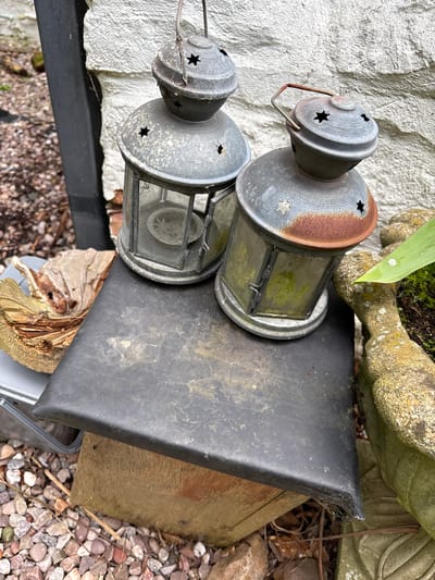 Two metal lanterns with star-shaped cutouts are stacked on a black plastic tray, which rests on a weathered wooden board and a piece of decaying wood. The scene is set against a backdrop of a white, textured wall, a stone planter with a green leaf, and a gravel-covered ground. Cobwebs are visible on the black plastic tray. The location is Pulverbatch, United Kingdom.