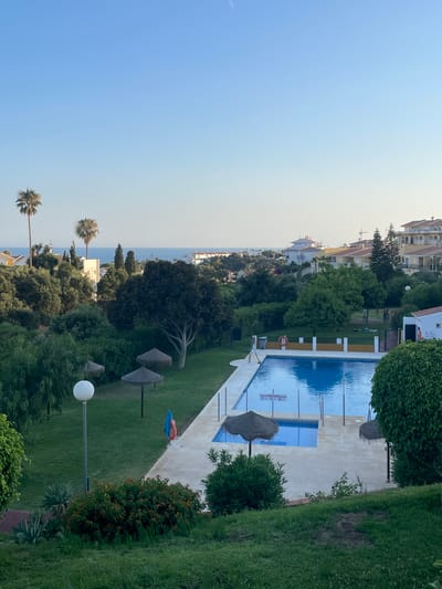 The image shows a view of a swimming pool area in Mijas, Spain, with the Mediterranean Sea in the distance. The pool is rectangular and filled with blue water. Around the pool, there is a tiled deck with several thatched umbrellas providing shade. In the foreground, there is a grassy lawn with some bushes and a large lamp post. In the background, there are trees, buildings, and palm trees, characteristic of the Mijas landscape. The sky is clear and blue, indicating a sunny day. There are no people clearly visible in the pool area, but a few figures can be seen in the distance on a grassy area near some buildings. The overall scene depicts a serene and sunny resort setting.