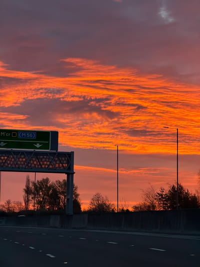 The image shows a sunset view from what appears to be a highway or motorway in the United Kingdom. The sky is filled with vivid orange and pink clouds. On the right side of the image, the dark asphalt of the road is visible, marked with white lane dividers. A grey concrete barrier separates the road from the roadside vegetation, which includes bare trees. Lamp posts stand along the roadside. A green road sign with white lettering directs traffic towards the M56 motorway. Based on the road sign indicating the M56 motorway, this photo was likely taken near Manchester, not Garden City, United Kingdom. There are no people visible.