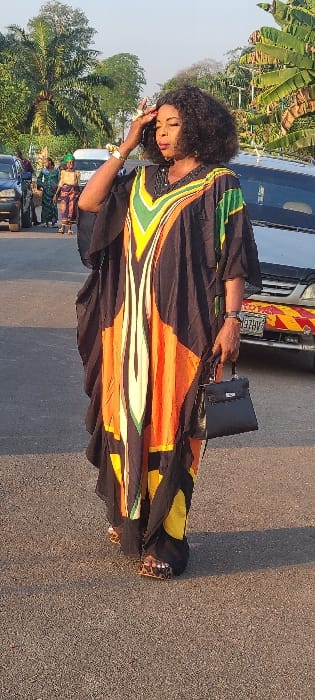 This image depicts an outdoor scene in Alayi, Nigeria, on a paved surface resembling a road or open area, under bright daylight. A middle-aged woman stands slightly off-center to the right in the foreground. She has dark, curly hair and wears a long, flowing kaftan-style dress that is primarily black with abstract vertical patterns in orange, yellow, and green, featuring a V-neck design. Her right hand is raised, with fingers touching her forehead, and she wears a golden bracelet and a prominent golden ring on her right hand. She carries a small, black, structured handbag with a silver-toned clasp in her left hand. Her feet are visible, wearing sandals with a leopard-print pattern.
In the background on the left, several individuals, predominantly women dressed in colorful traditional attire and head wraps, are visible. Behind them, a dark-colored SUV is partially present. Further back, numerous palm trees and other green foliage are visible. On the right side of the background, a silver-colored vehicle, identified as a Toyota by a visible logo, is partially seen, displaying a license plate containing "AHZE" and a yellow and red striped reflective sticker on its lower bumper. Additional green trees, including what appear to be banana trees, are present on the far right. The ground surface is dark gray asphalt.