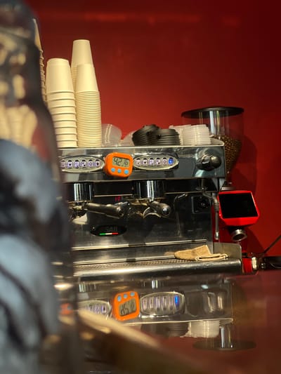 The image shows a close-up, eye-level view of a chrome espresso machine against a bright red wall. Several stacks of white paper cups are visible on top of the machine, along with clear plastic lids and a black cap. A glass coffee bean grinder is positioned behind the cups. On the front panel of the espresso machine, a small orange digital timer displays "42.16". To the right of the timer is a red screen with a black display. A black coffee portafilter is attached to the machine, and a small brown cloth rests on the drip tray. The reflection of the machine and cups is visible on the shiny countertop below. The image appears to be taken in a commercial setting, likely a cafe or coffee shop, possibly located in Ochamchire, Abkhazia.