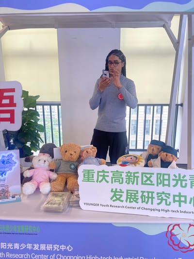 In an indoor setting, possibly a booth at an exhibition or event, a person with dark skin and braided hair wearing glasses and a grey long-sleeved shirt is holding a smartphone and looking at it. The person is standing behind a table displaying several teddy bears and some merchandise. To the left of the person is a tall green plant. On the table, there are at least five teddy bears of various sizes and colors, some wearing clothes. In front of the teddy bears, there are stacks of what appear to be trading cards or small brochures. A banner with Chinese characters in green and white is displayed behind the teddy bears. The banner reads "重庆高新区阳光青少年发展研究中心" and "YOUNGER Youth Research Center of Chongqing High-tech Industrial Development Zone". Another banner with similar Chinese characters and the English translation "ith Research Center of Choanging High-tech Industrial Development Zone" is visible at the bottom of the image, suggesting this event is related to the Chongqing High-tech Industrial Development Zone. The background shows a window with buildings visible outside, indicating an urban environment. The overall impression is of a promotional event or a display for a youth research center. The location is identified as 龙溪街道, China, which is a subdistrict within Chongqing.