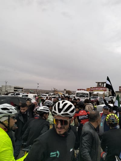 A crowd of cyclists, mostly wearing helmets and cycling gear, gathers in a parking lot. Many wear casual attire. Some hold black and white flags. A white van and multiple cars are parked behind the crowd. Buildings are visible in the background under an overcast sky. The cyclists are likely at the starting point of a race.